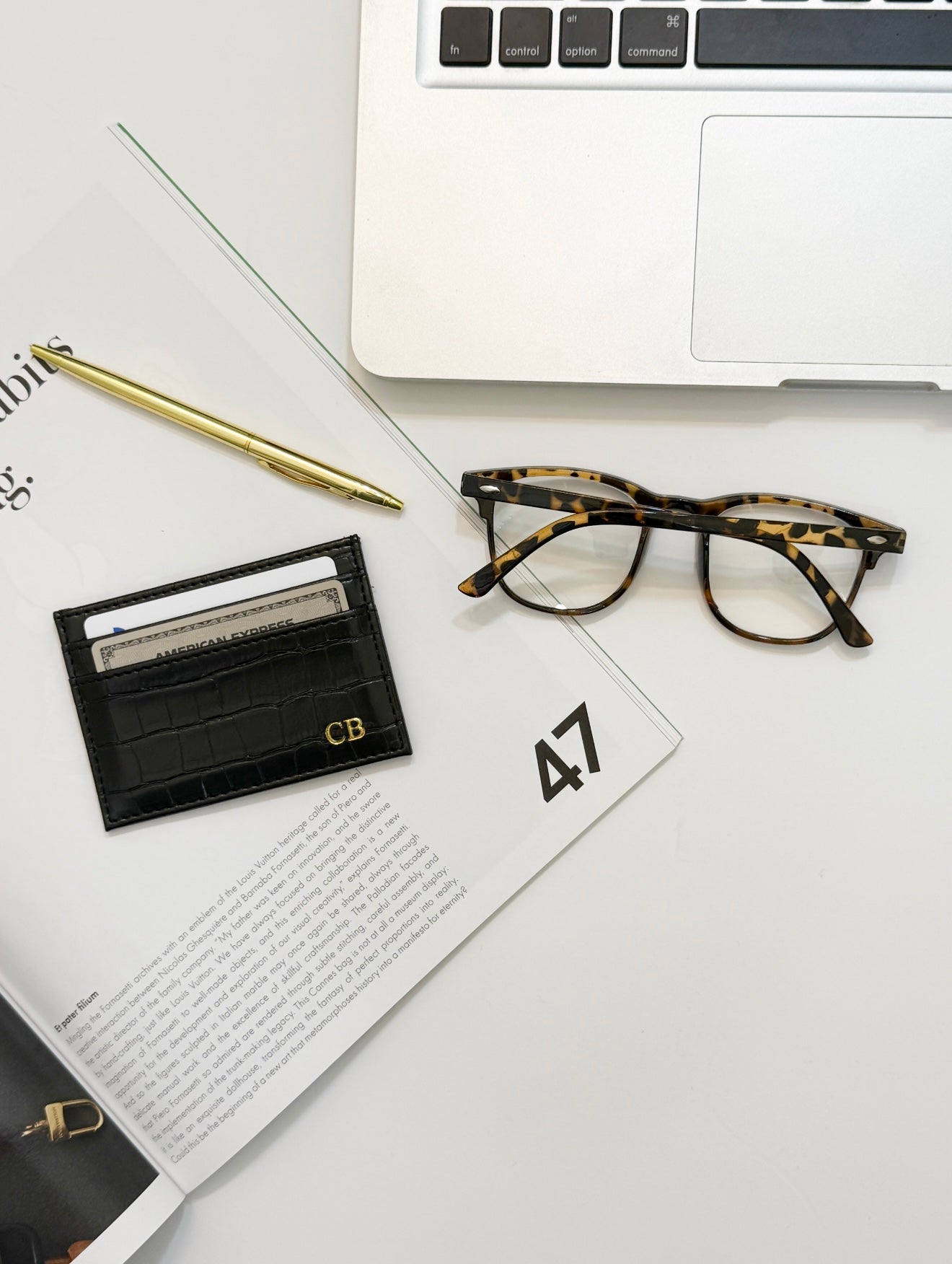 Black crocodile vegan leather cardholder laying beside a gold pen,  reading glasses, and laptop