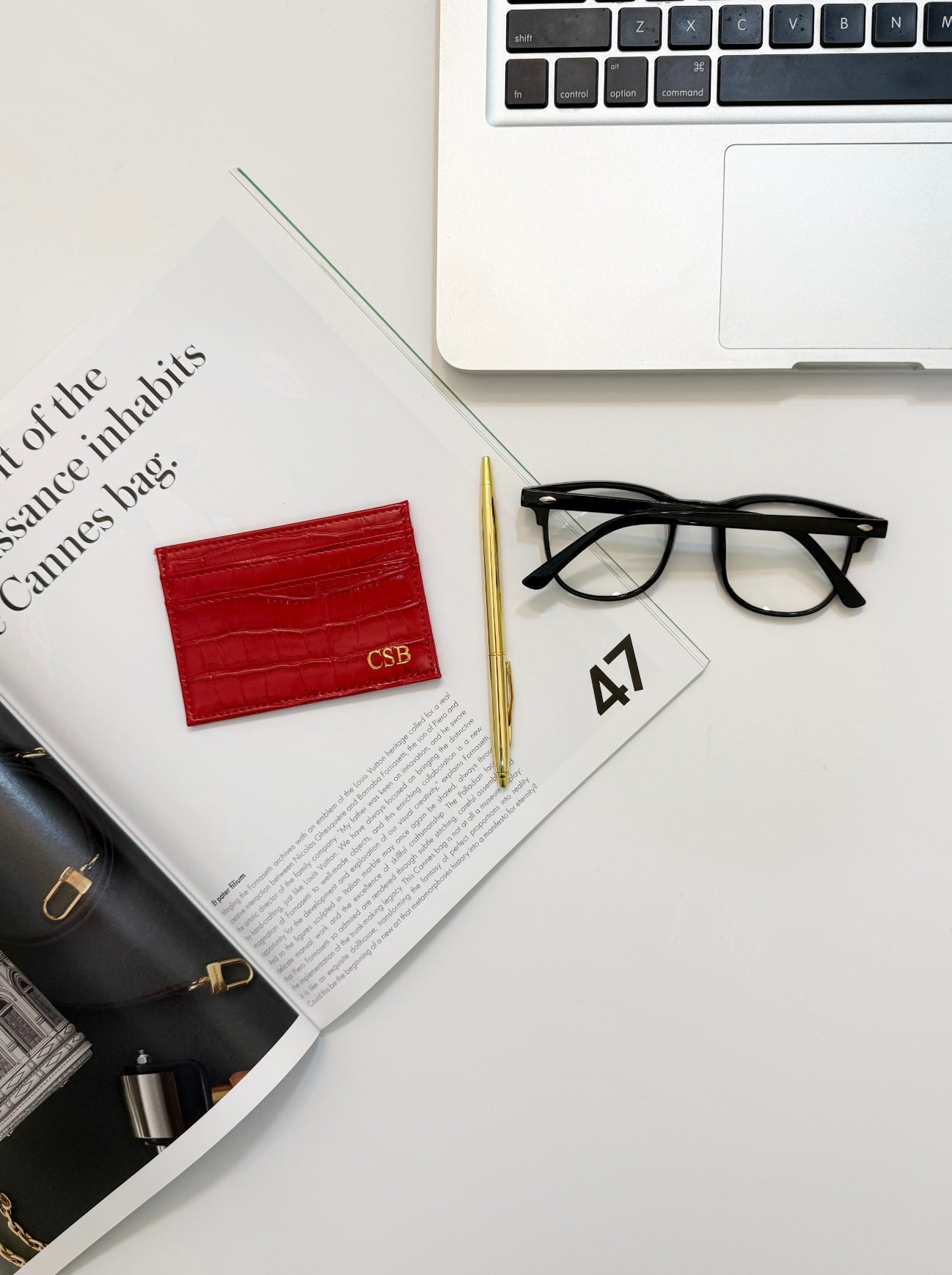 Monogrammed red vegan crocodile leather cardholder beside a gold pen, reading glasses, and a laptop.