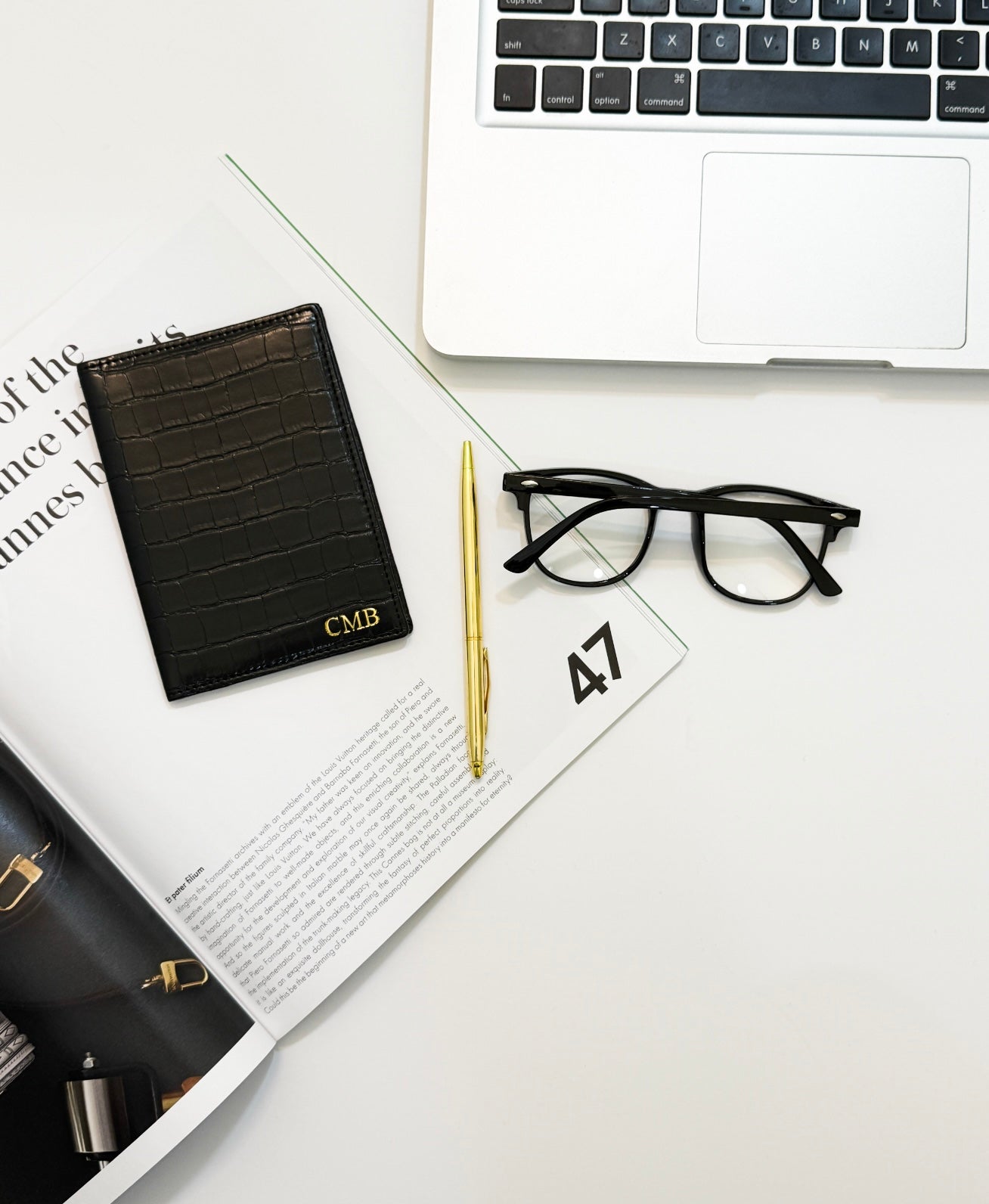 Black monogrammed passport cover by a gold pen, reading glasses, and a laptop 