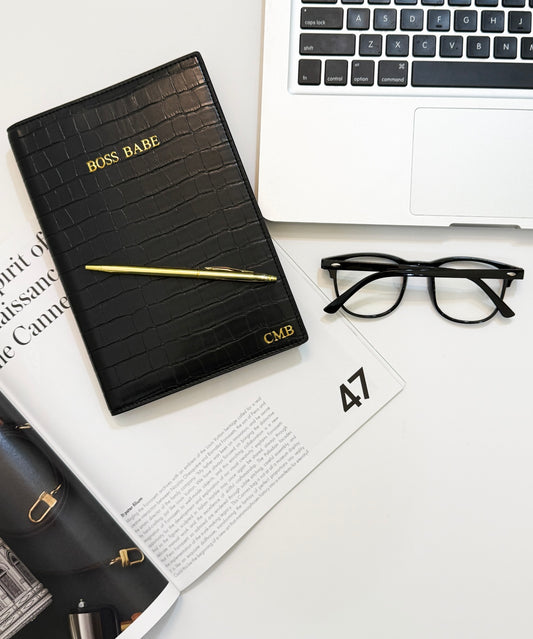 Black vegan crocodile monogrammed  notebook  cover with "boss babe" written on front. This cover is beside a gold pen, reading glasses, and a laptop.  