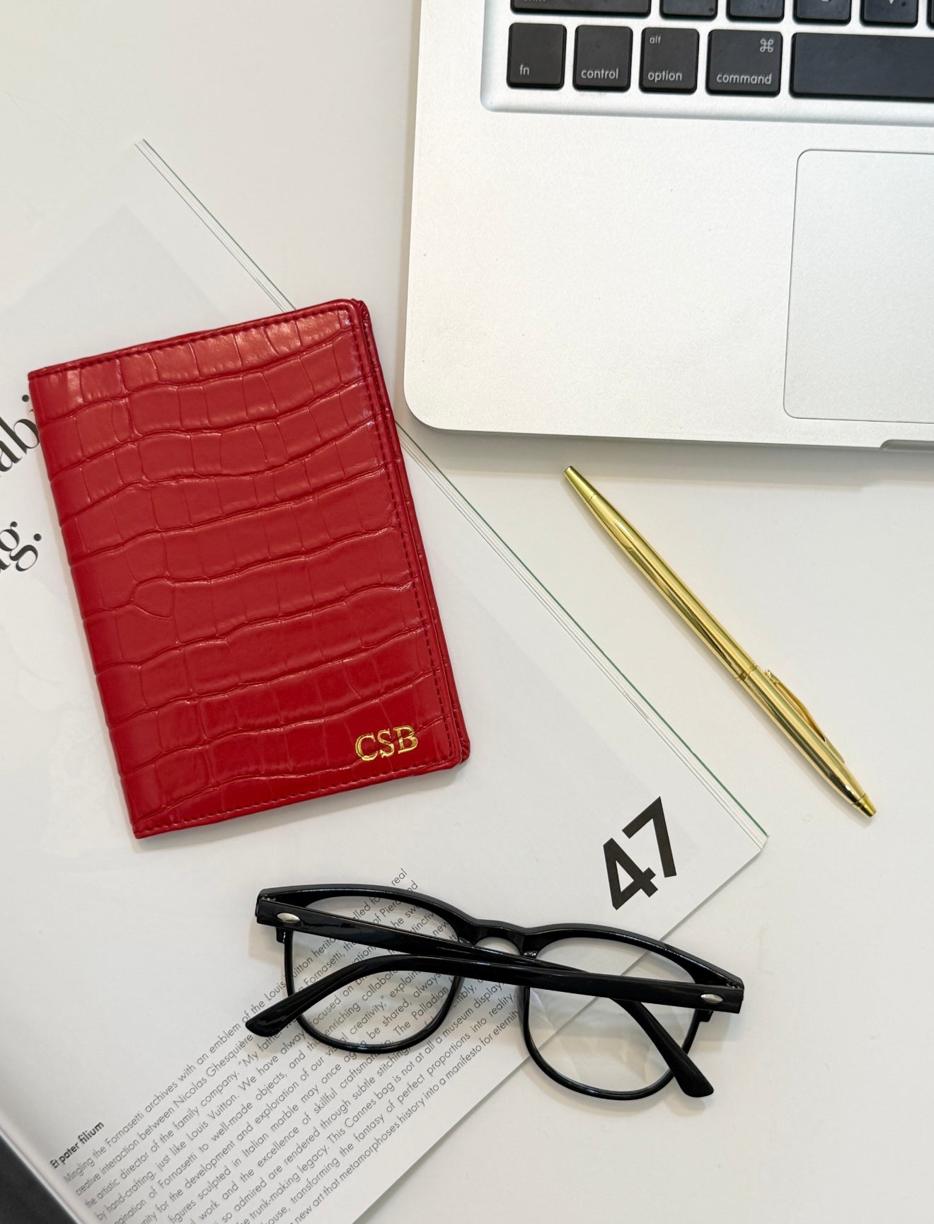 Red monogrammed  crocodile passport cover beside reading glasses, a gold pen, and a laptop. 